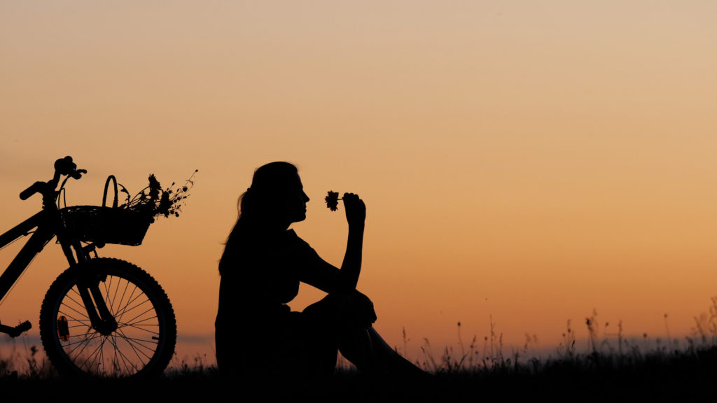 young woman silhouette, being present in the moment, sitting next to a bike on hill and smelling a flower