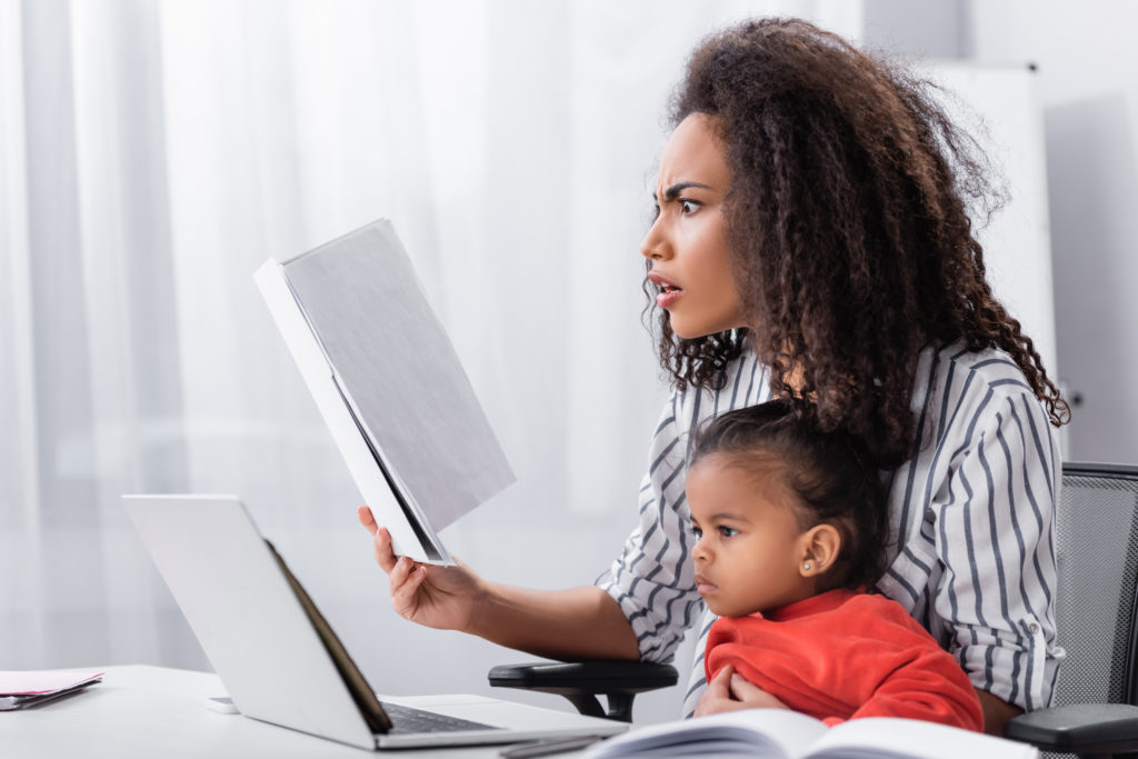 mother stressed from a lack of self-care holding notes while sitting with toddler daughter and working from home